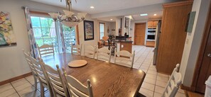 A dining area with a wooden table and chairs, a chandelier, and a view of the backyard through large windows.