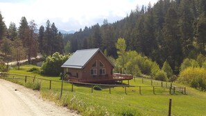 A rustic cabin with a metal roof, surrounded by a wooden deck and a fenced yard, set in a forested valley.