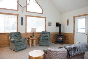A cozy cabin interior with two armchairs, a small table, a round wooden table, a sofa, a fireplace, and a wall-mounted deer skull.