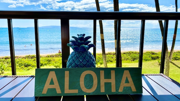 Eine Holzterrasse mit einem Schild mit der Aufschrift 'ALOHA' und einer Ananasdekoration, mit Blick auf einen Strand bei klarem Himmel und Meerblick.