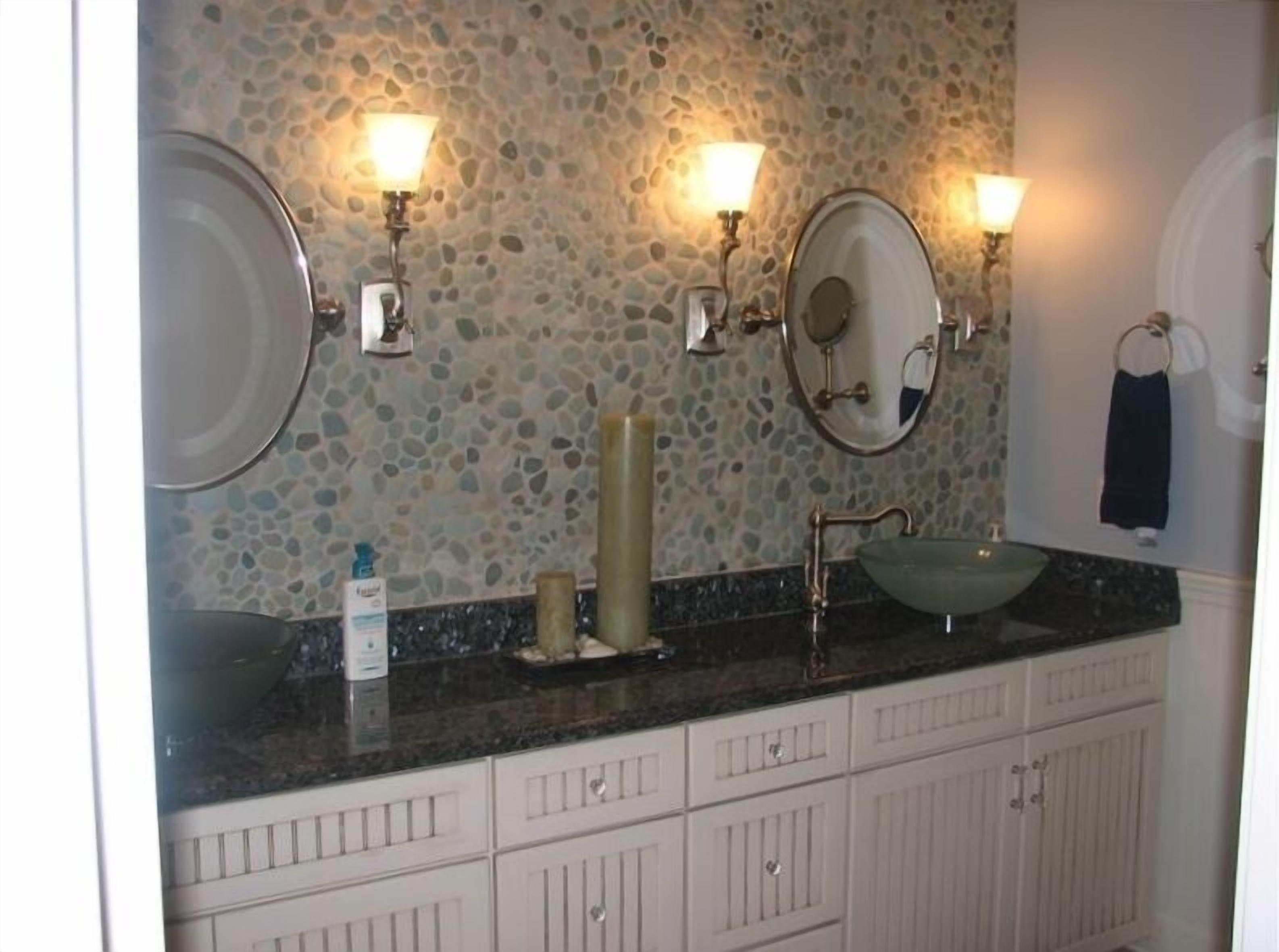 A bathroom with a stone mosaic wall, two wall-mounted lights, a round mirror, a sink with a green basin, and a white cabinet with drawers.