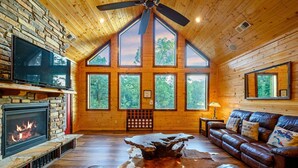L’intérieur d’une cabane en rondins, avec une cheminée en pierre, un canapé en cuir et de grandes fenêtres donnant sur la forêt.