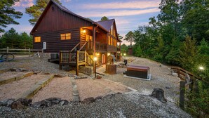 Un chalet rustique en rondins de bois, doté d’un porche et d’un jacuzzi, entouré d’une allée en gravier et d’une zone boisée.