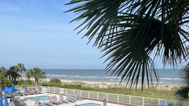 A pool area with lounge chairs and umbrellas, surrounded by a white fence, overlooking a beach.