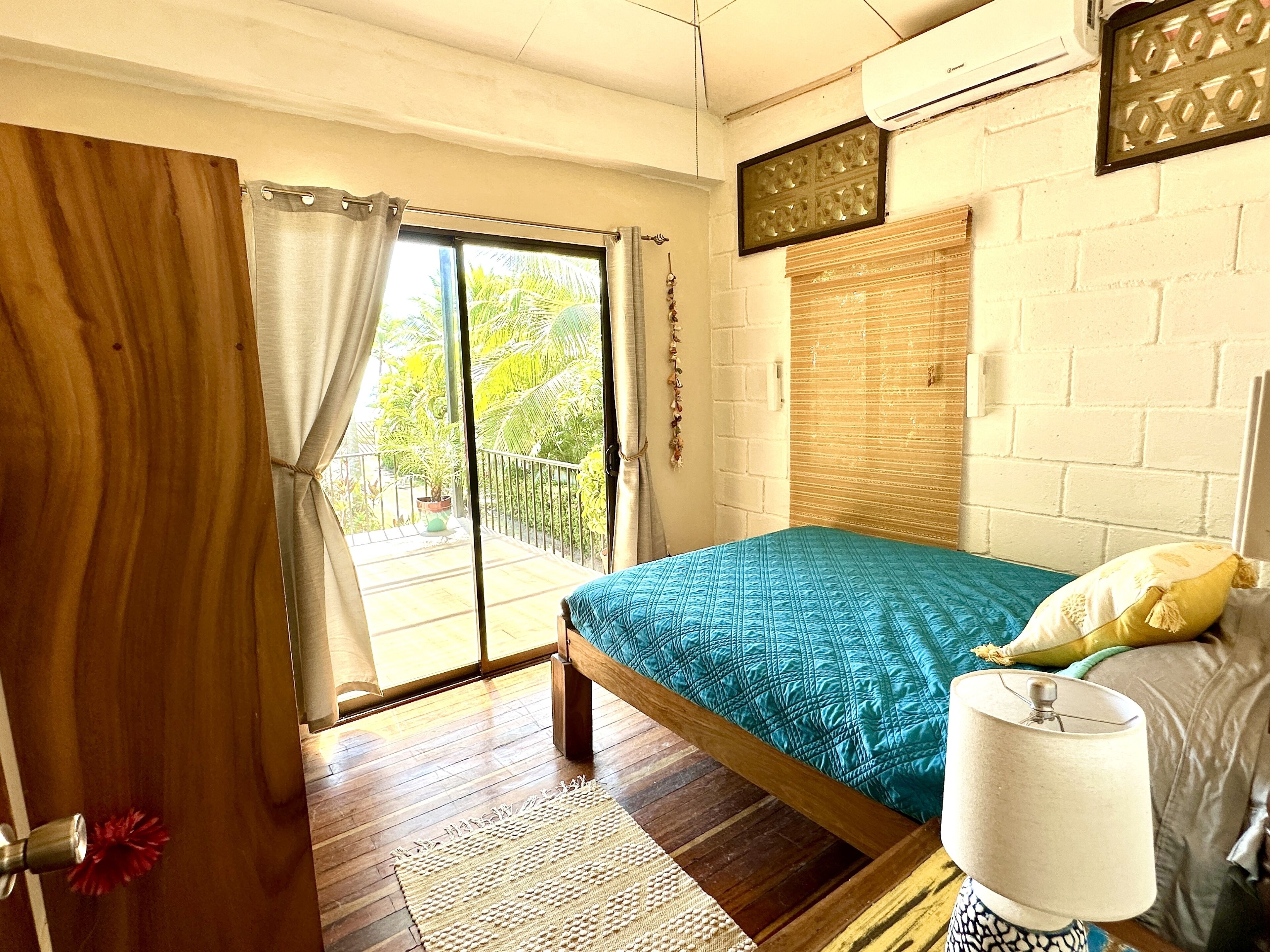 A bedroom with a bed, a window with curtains, a wooden floor, and a wall-mounted air conditioner.