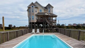 Eine Holzterrasse mit einem rechteckigen Swimmingpool, eine Holztreppe, die zu einem Haus mit mehreren Fenstern und einem Balkon führt.