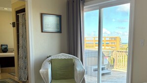 Ein Zimmer mit einem Bett, einem Korbsessel mit grüner Sitzkissen, einem gerahmten Bild an der Wand und einer Glastür zu einem Balkon mit Blick auf Gebäude und eine Holzterrasse.