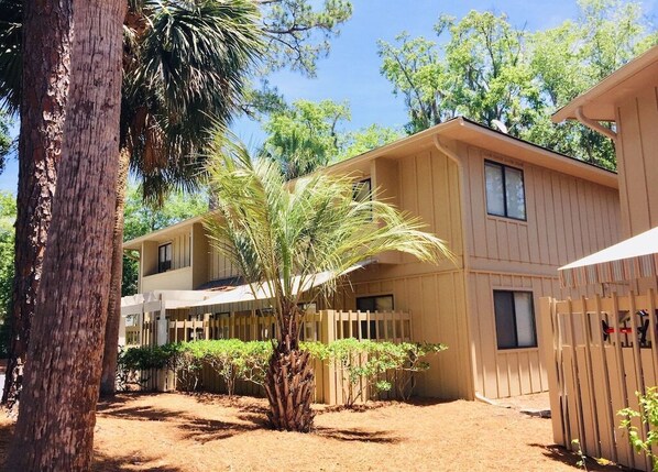 A two-story building with a wooden exterior, a fenced patio area, and a palm tree. - Having fun in the Sun Beach Villa (Hilton Head Island)