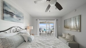 Un dormitorio con cama, ventilador de techo, vista a un área exterior con mesa y sillas, y obras de arte en las paredes.