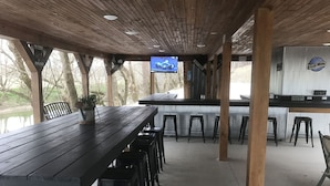A covered outdoor seating area with a long table and multiple stools, a television mounted on the wall, and a sign indicating a parking rate. - Cozy Cabin center of Bourbon Trail, Near Bardstown, beautiful country views (Springfield)
