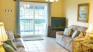 A living room with a sofa, a television, a ceiling fan, and a large window with curtains.