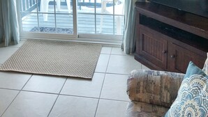 A living room with a flat-screen TV on a wooden cabinet, a patterned sofa, a woven rug, and a sliding glass door leading to a balcony with outdoor furniture.