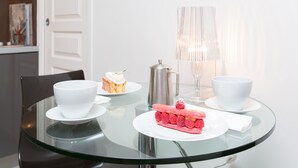 Une table à plateau en verre, avec des tasses blanches, une cafetière, un dessert et une lampe.