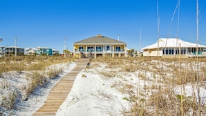 Une maison de plage dotée d’une terrasse en bois menant à l’entrée, entourée de sable et d’une végétation clairsemée.