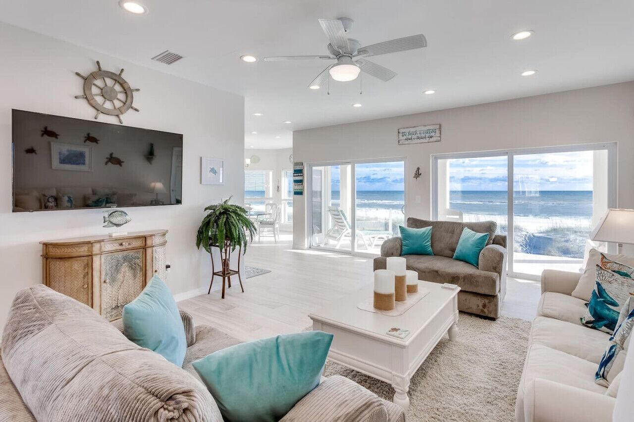 A spacious living room with a large TV, comfortable seating, a ceiling fan, and a view of the ocean.