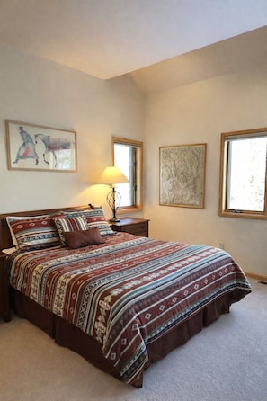 A bedroom with a large bed, two paintings, a nightstand with a lamp, and a window.