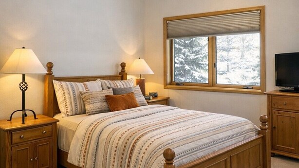 A bedroom with a wooden bed, two bedside tables with lamps, a television, and a window with a view of trees. - Ski-in, Quick Walk to Slopes with AMAZING VIEWS (Mt Crested Butte)