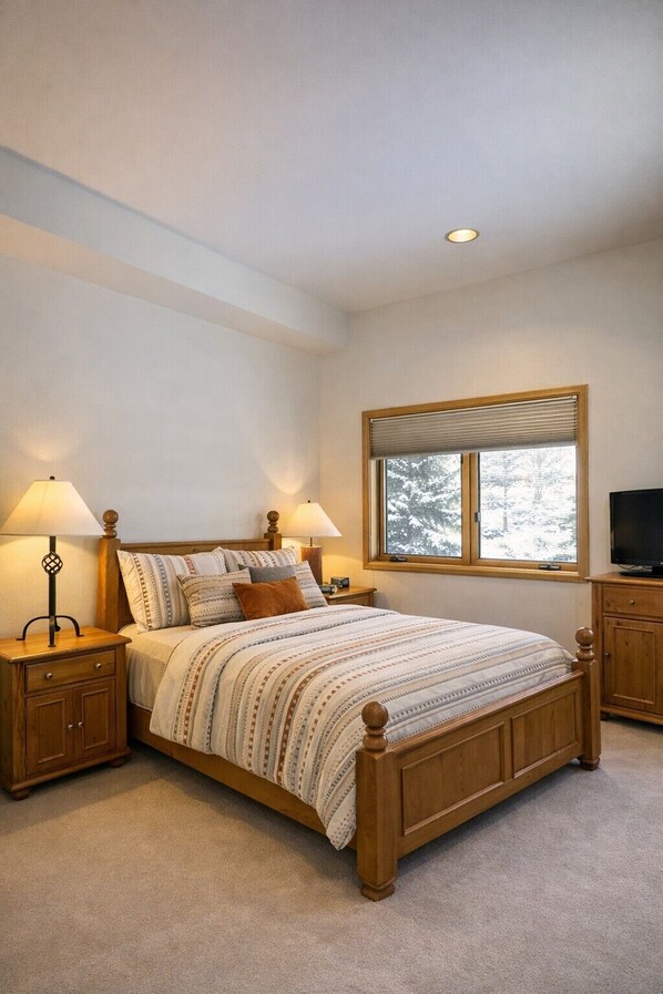 A bedroom with a wooden bed, two bedside tables with lamps, a television, and a window with a view of trees.