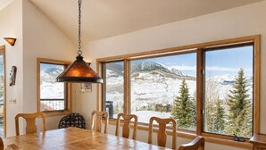 A dining area with a wooden table and chairs, large windows with a snowy mountain view, and a hanging pendant light. - Ski-in, Quick Walk to Slopes with AMAZING VIEWS (Mt Crested Butte)