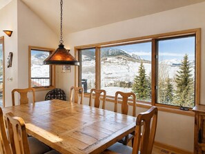 A dining area with a wooden table and chairs, large windows with a snowy mountain view, and a hanging pendant light. - Ski-in, Quick Walk to Slopes with AMAZING VIEWS (Mt Crested Butte)