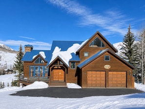 A large wooden cabin with a garage, a covered porch, and a snow-covered landscape.