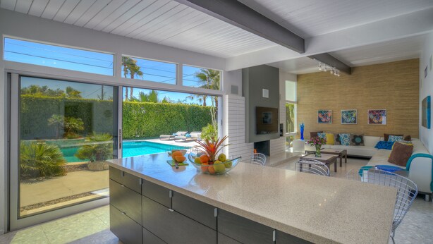 A modern kitchen with a large island, a dining area with a sofa and table, and a view of a swimming pool and garden. - PS Butterfly: Well-Styled, Authentic Mid-Century Home (Palm Springs)