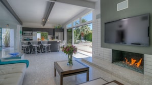 A modern living room with a fireplace, a TV mounted on the wall, and a kitchen in the background. - PS Butterfly: Well-Styled, Authentic Mid-Century Home (Palm Springs)