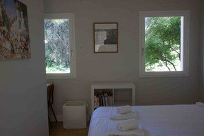 A bedroom with a bed, a small white cabinet with books, two windows, and a painting on the wall. - Stunning and spacious home in Provence! (Lourmarin)