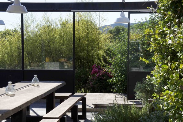 An outdoor patio with a wooden table and benches, surrounded by lush greenery and a view of distant hills. - Stunning and spacious home in Provence! (Lourmarin)