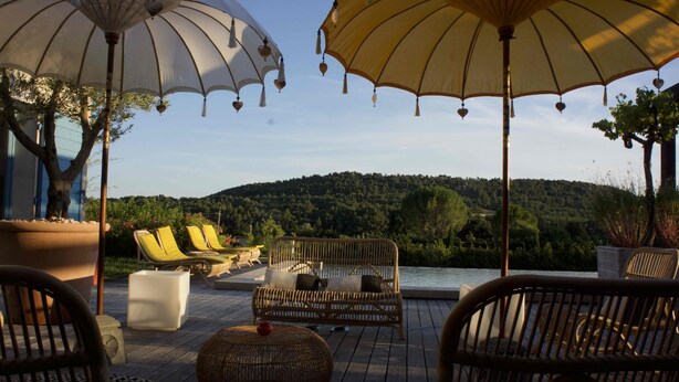 Eine Terrasse mit Korbmöbeln, Sonnenschirmen und Blick auf einen bewaldeten Hügel.