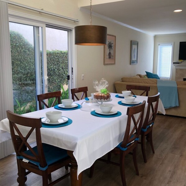 Comedor con una mesa dispuesta para seis personas, sillas, lámpara colgante y vista a follaje exterior.