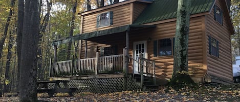 Une cabane en rondins dotée d’un porche et d’une terrasse, entourée d’arbres et de feuilles mortes.