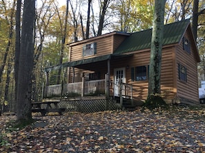 Une cabane en rondins dotée d’un porche et d’une terrasse, entourée d’arbres et de feuilles mortes.