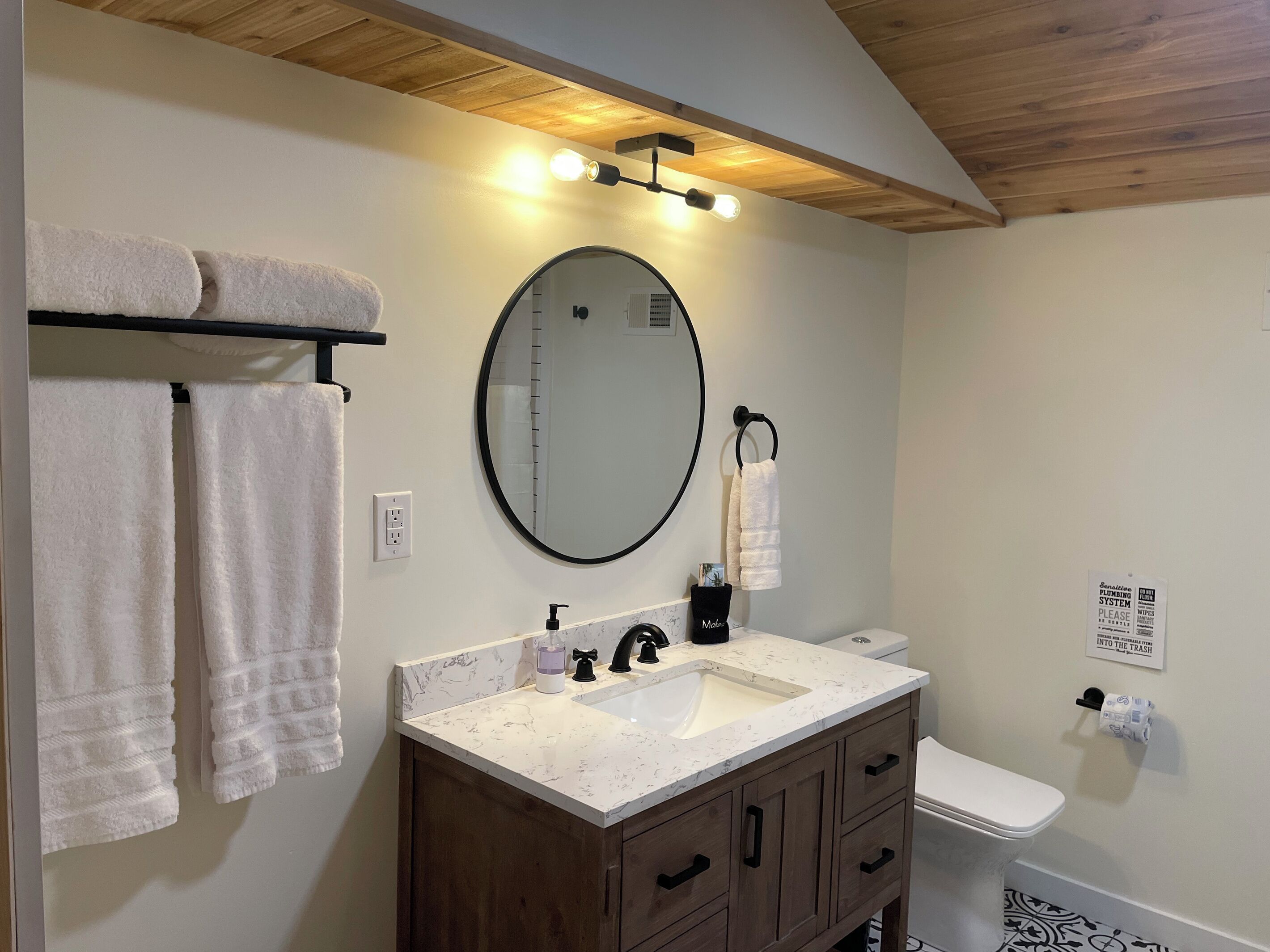 Une salle de bain avec un plafond en bois, un miroir rond, un lavabo avec un plan de toilette en marbre, des toilettes et deux serviettes suspendues à un porte-serviettes.