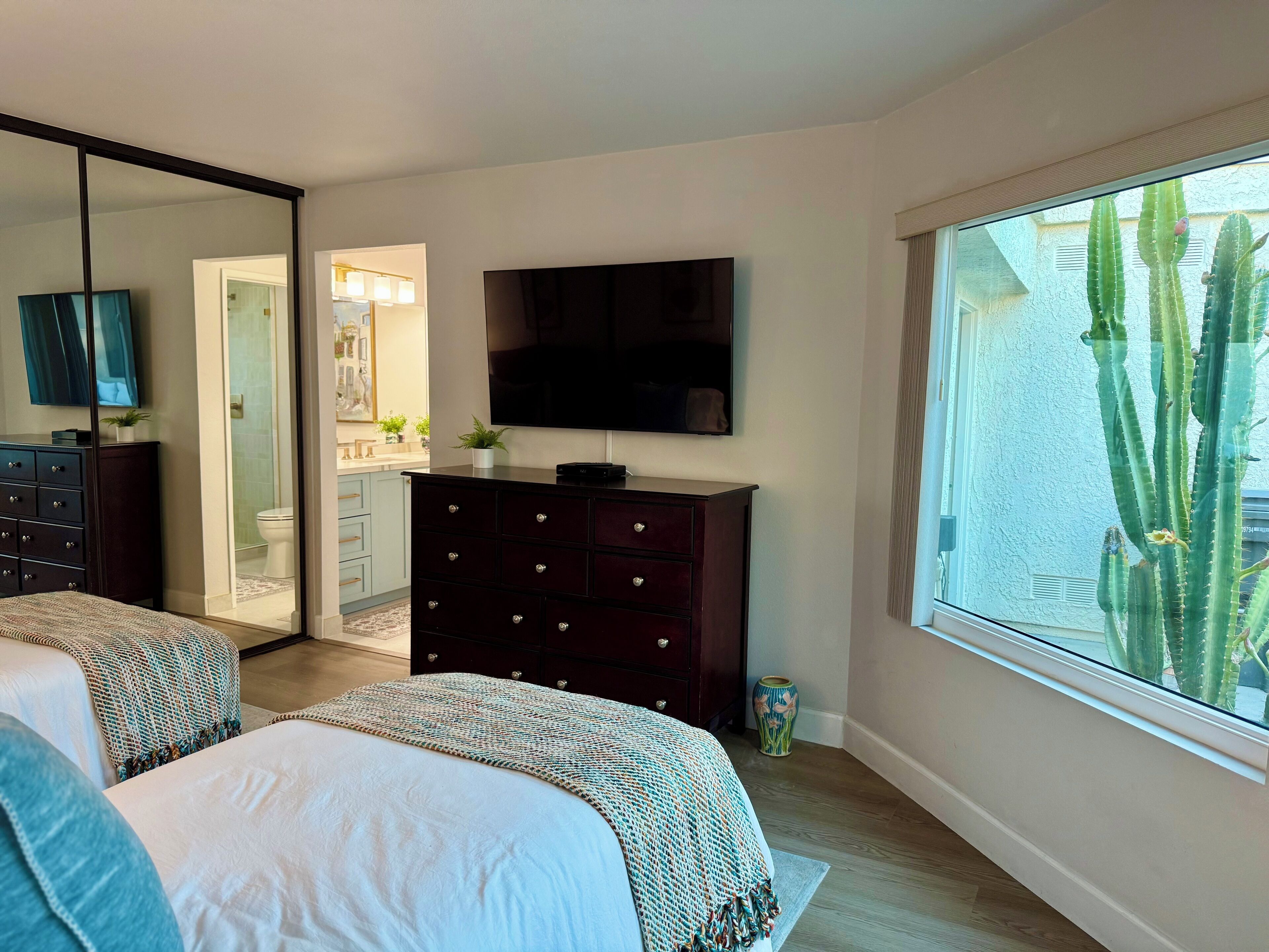 A bedroom with a bed, a dresser, a TV, and a large window with a view of cacti.