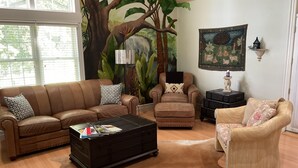 Una sala de estar con un sofá de cuero marrón, un sillón marrón, una mesita redonda y una mesa de centro. En la pared detrás del sofá, hay un mural grande de un árbol y una selva.