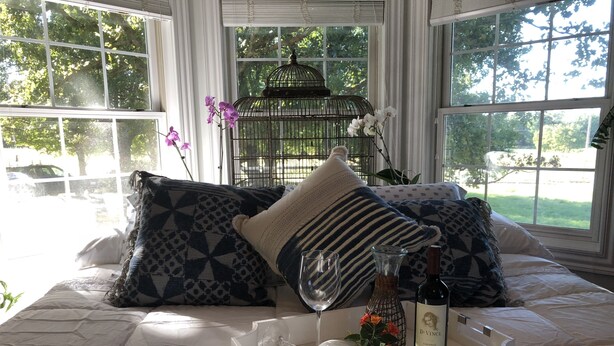 Una cama con almohadas estampadas, una bandeja con una botella de vino y copas, y una jaula decorada con flores.