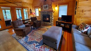 A cozy log cabin interior with a stone fireplace, wooden walls, and a large window. - McKenzie Riverfront Cabin big screened-in porch, 1/2mi to Mckenzie River Trail (McKenzie Bridge)