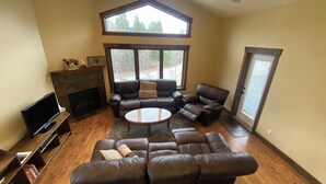 A spacious living room with a fireplace, leather sofas, a television, and a large window offering a view of trees. - Luxury Walk Out House in Radium Hot Springs 5 Bedrooms (Radium Hot Springs)