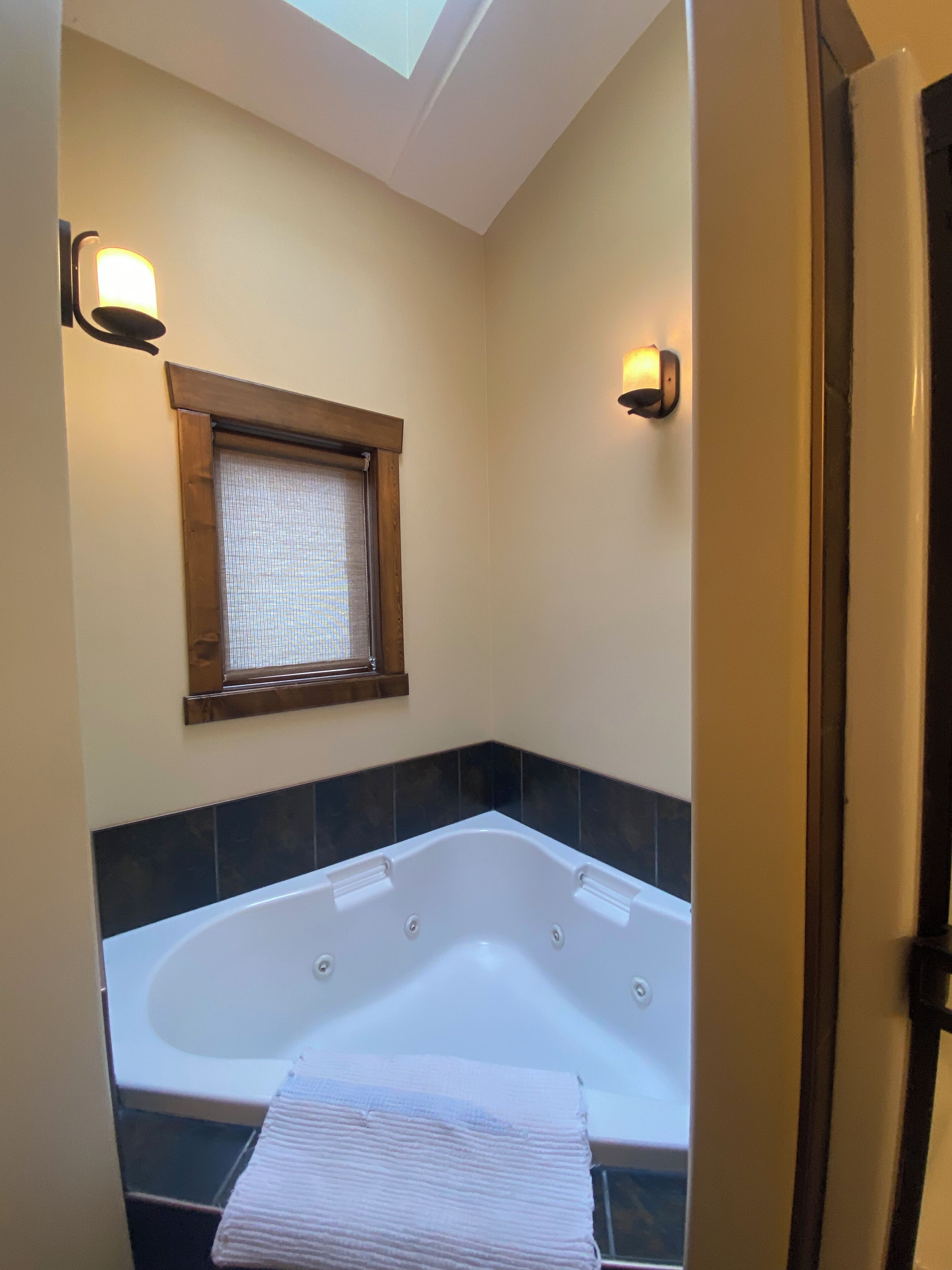 A bathroom with a large bathtub, a window with blinds, and two wall-mounted lights.
