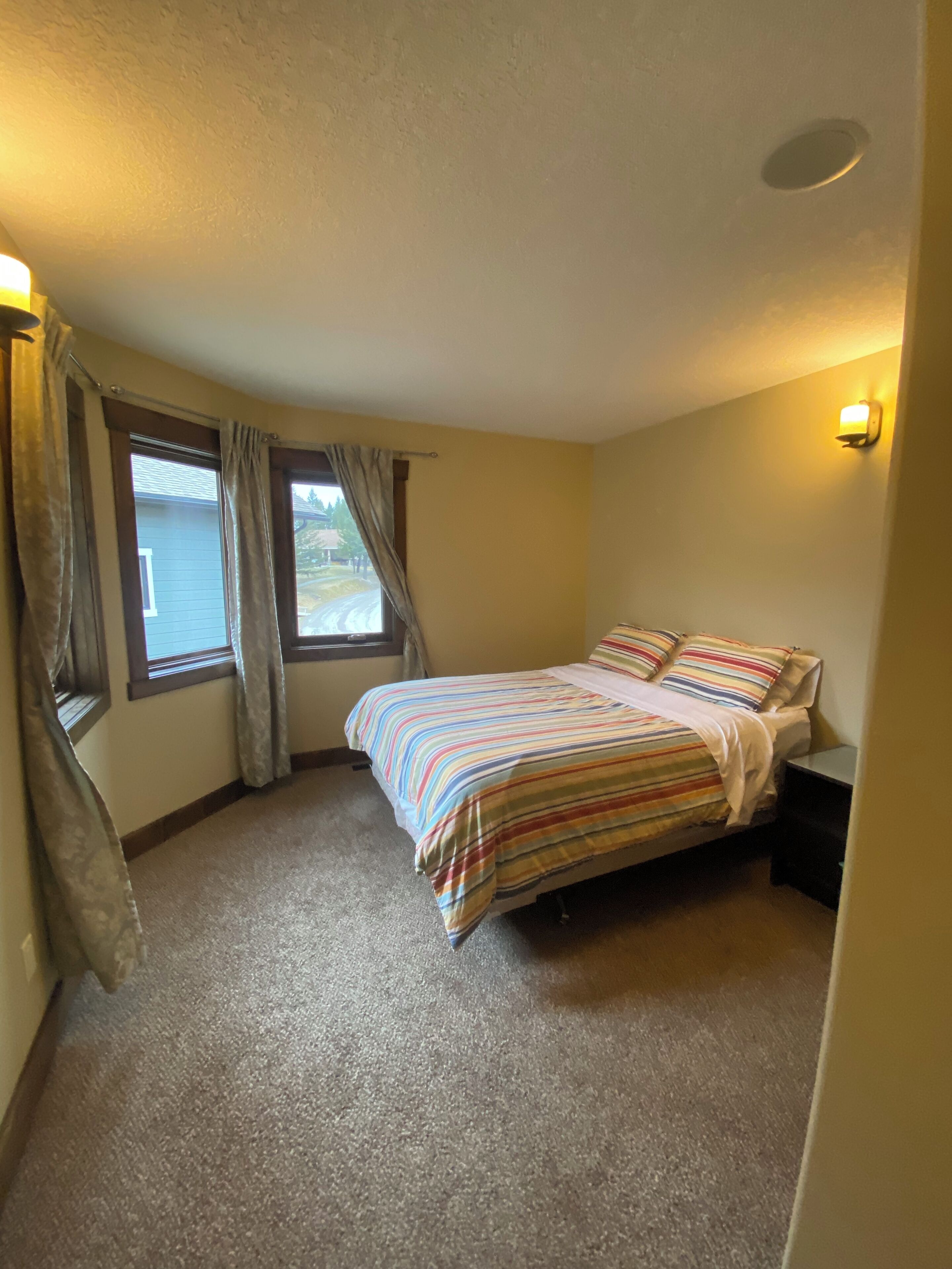 A bedroom with a bed, a nightstand, a window with curtains, and a wall-mounted light.