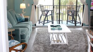 A living room with teal walls, a ceiling fan, a white coffee table, a grey rug, and a balcony with chairs. - 2 Bedroom Ocean View Condo, walk to restaurants/bars, 2 pools on premises (St Augustine)
