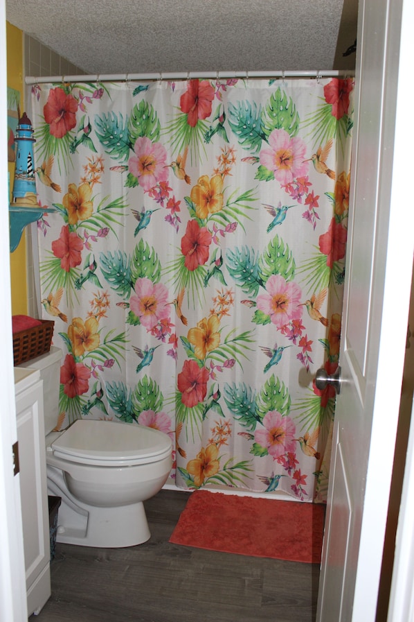 A bathroom with a toilet, a shower curtain with a floral pattern, and a red bath mat.