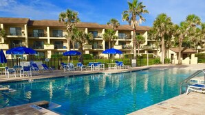 A swimming pool with blue chairs and umbrellas, surrounded by a building with balconies and palm trees. - 2 Bedroom Ocean View Condo, walk to restaurants/bars, 2 pools on premises (St Augustine)