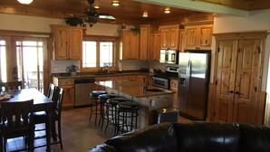 A spacious kitchen with wooden cabinets, a central island with stools, a stainless steel refrigerator, and a dining area with a leather couch. - Remote ranch house on 93 acres in the peaceful Indianola Valley! Sleeps 30 (Fairview)
