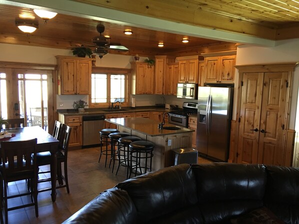 A spacious kitchen with wooden cabinets, a central island with stools, a stainless steel refrigerator, and a dining area with a leather couch. - Remote ranch house on 93 acres in the peaceful Indianola Valley! Sleeps 30 (Fairview)