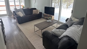 Een ruime woonkamer met een donkerbruine leren bank, een bijpassende fauteuil en een salontafel. Een flatscreen televisie is aan de muur bevestigd, en er is een groot raam met uitzicht op de omgeving.