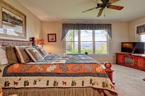 A bedroom with a large bed, a ceiling fan, a TV, and a window with a view of the outdoors.