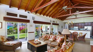 A spacious living room with a wooden ceiling, a large sofa set, a coffee table, wicker furniture, and a view of the outdoors through large glass doors.