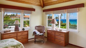 A room with a bed, two wicker side tables, a chair, and large windows with a view of the ocean and greenery.